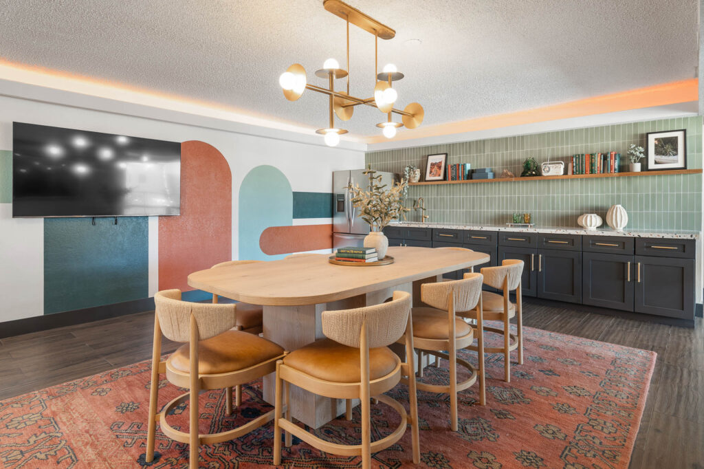 A modern dining area with a wooden table, six tan chairs, a colorful rug, wall art, a large flat-screen TV, and cabinets with books and decor under a geometric chandelier.