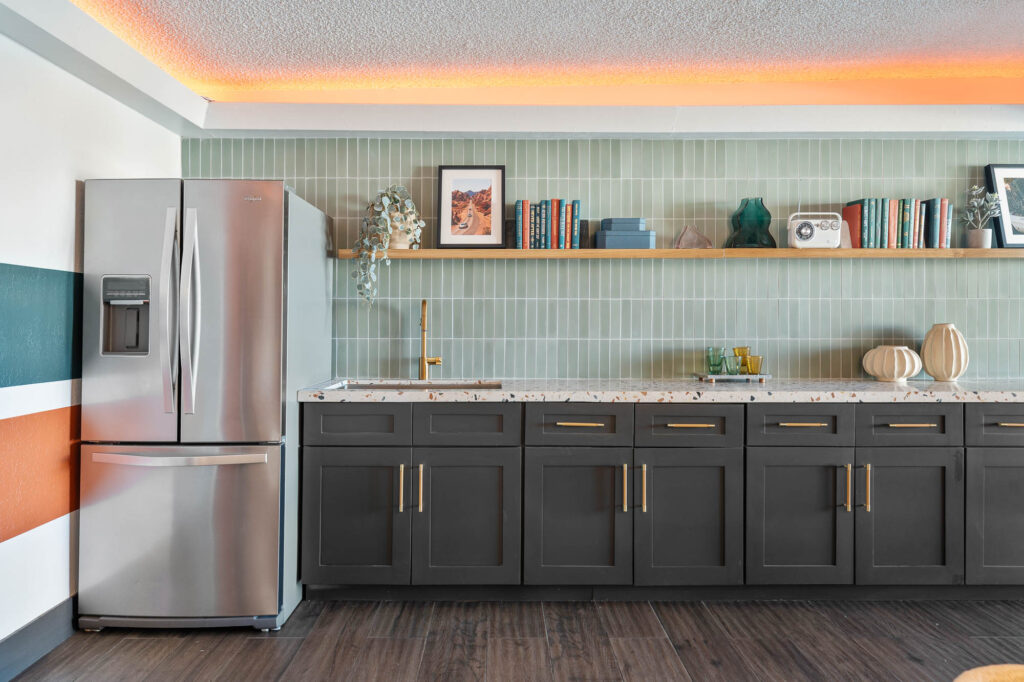 Modern kitchen with a stainless steel refrigerator, dark cabinets with gold handles, a long terrazzo countertop, green tiled backsplash, and a wooden shelf displaying books, decor, and a framed photo.