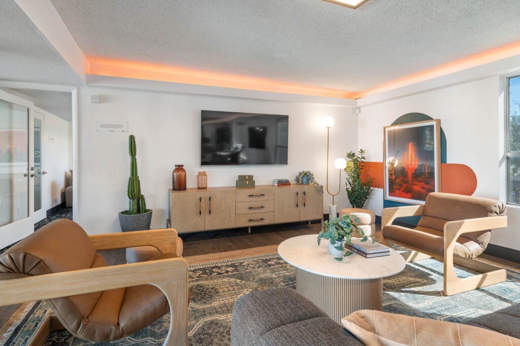 Modern living room with neutral decor, wooden armchairs, a round coffee table, potted plants, a large TV mounted on the wall, and artwork leaning against the wall. Warm lighting highlights the ceiling.
