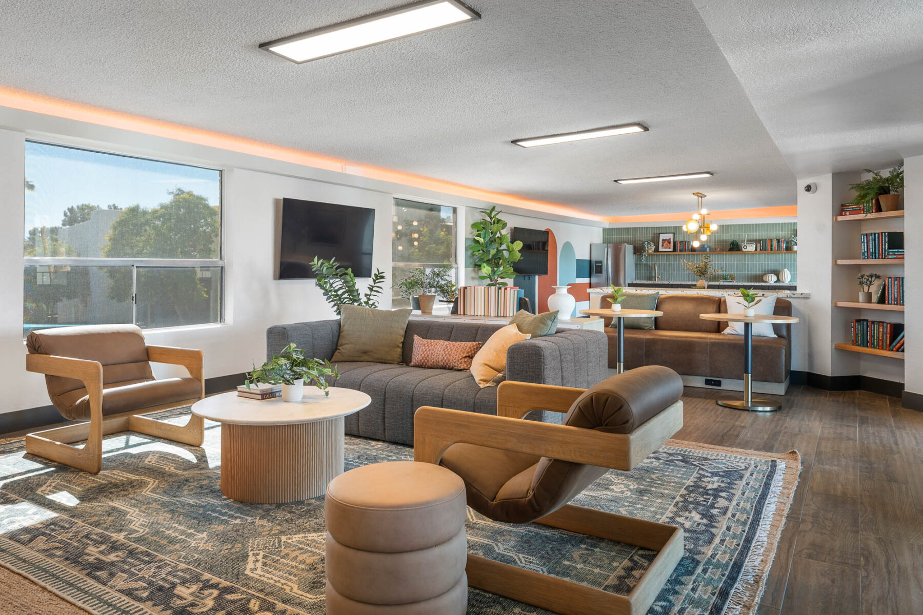 Modern living room with a gray sofa, wooden chairs, round coffee tables, stools, bookshelf, plants, and large windows letting in natural light. A kitchen and dining area are visible in the background.