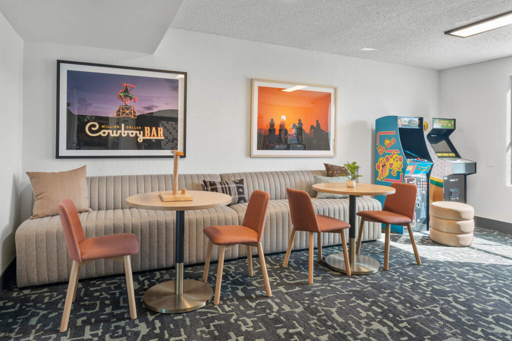 A lounge area with a striped sofa, round tables, orange chairs, framed art on the wall, two arcade machines, and natural light from a window on the right.