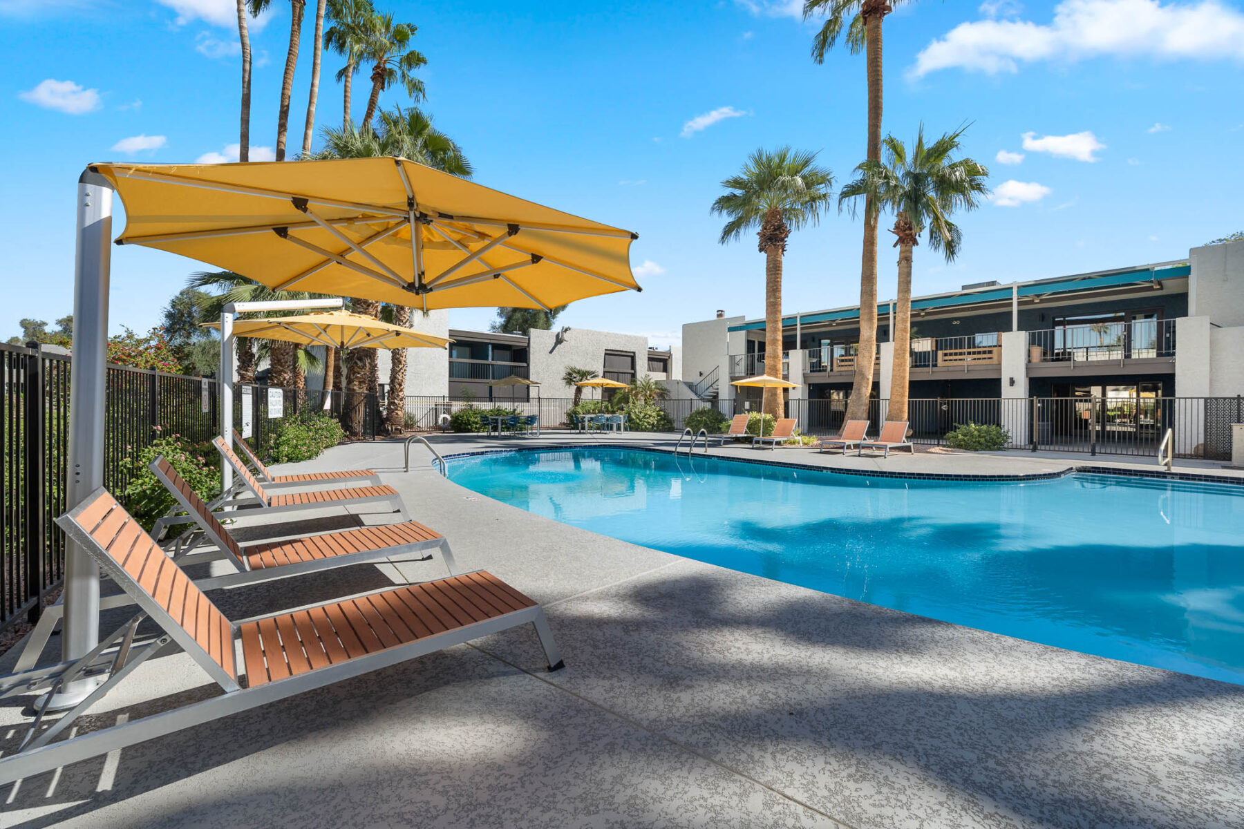 Outdoor swimming pool with clear blue water, surrounded by lounge chairs and large yellow umbrellas. Palm trees and modern apartment buildings are in the background under a sunny sky with scattered clouds.