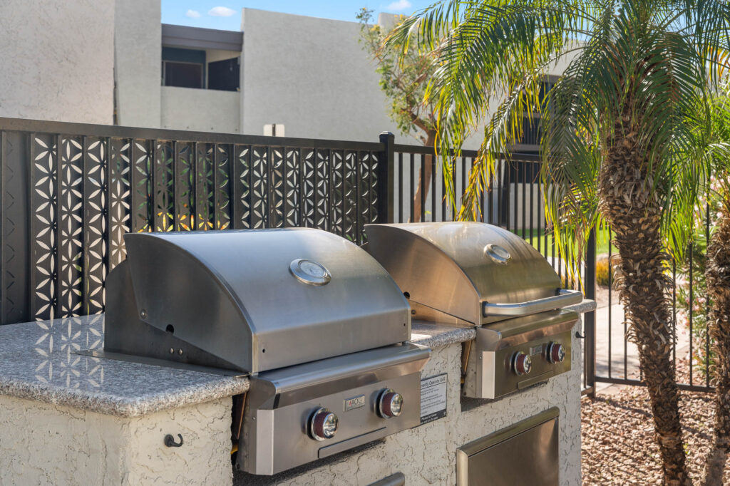Two stainless steel outdoor grills with closed lids are set on a stone countertop next to a decorative black metal fence and a palm tree, with modern white buildings in the background.