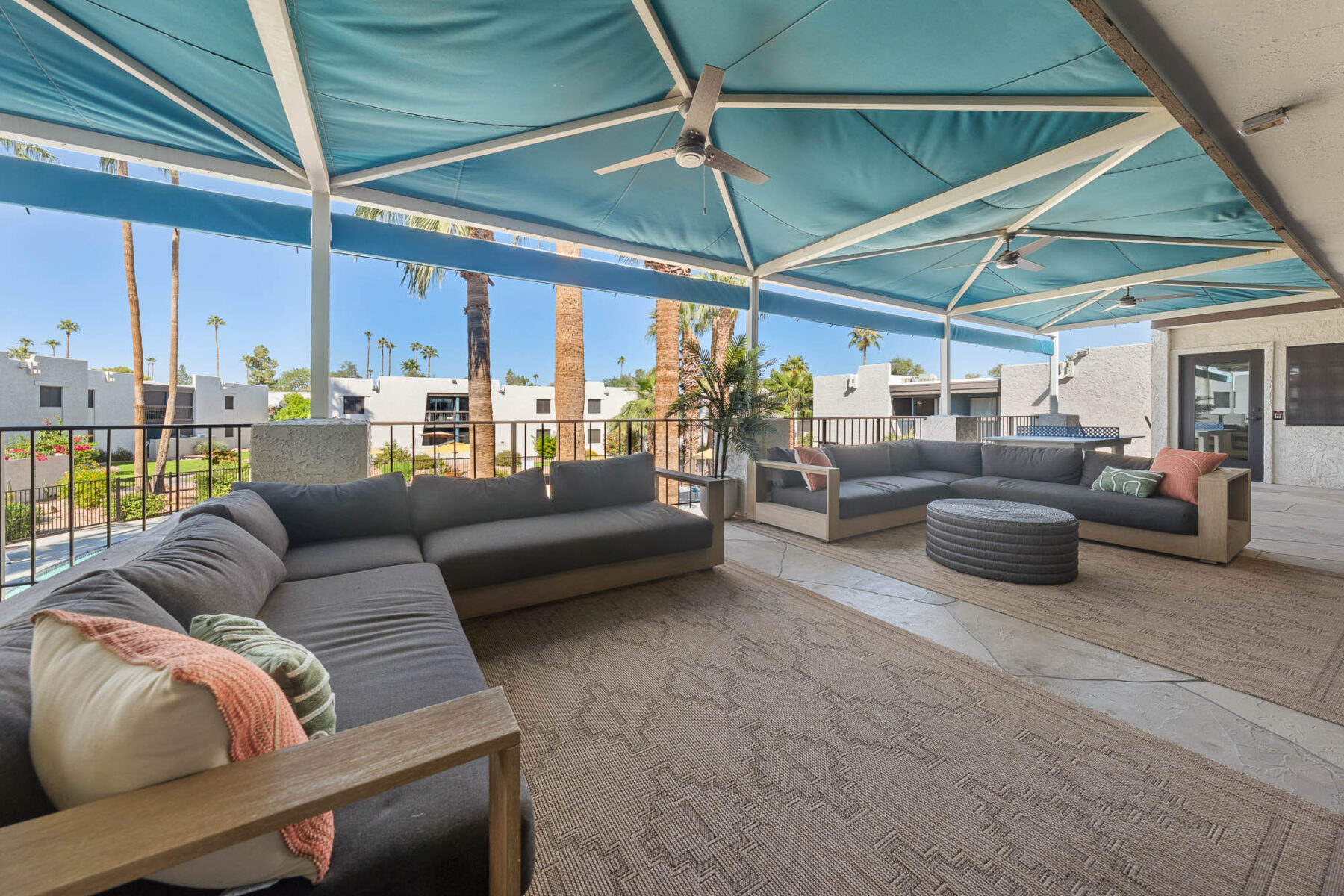 A covered outdoor lounge area with gray sectional sofas, cushions, a round ottoman, ceiling fans, and a large rug. The space overlooks a pool and palm trees, with modern buildings visible in the background.