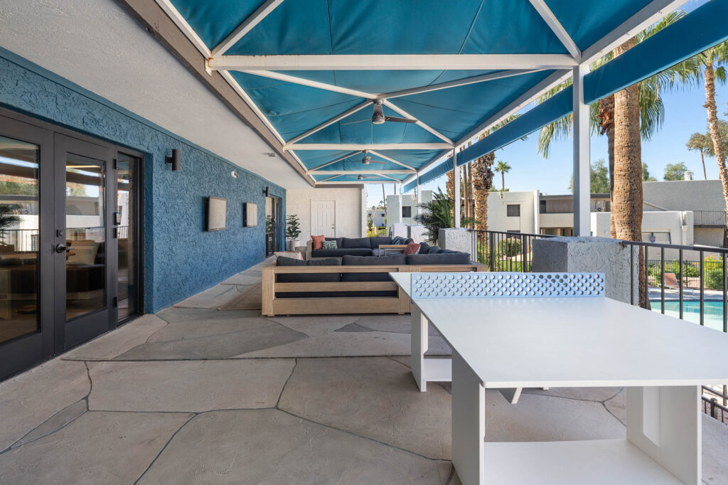 Outdoor lounge area with blue awning, modern seating, and a white ping pong table overlooking a pool and palm trees, next to a building with blue walls and large glass doors.