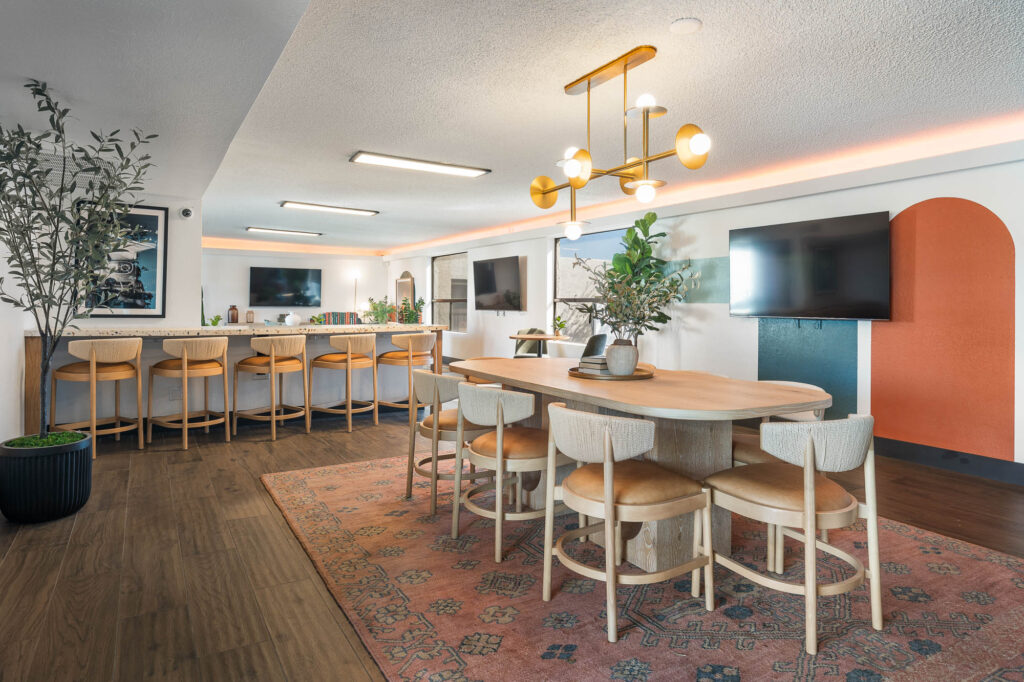 Modern communal dining area with a long wooden table, light chairs, bar stools at a counter, two wall-mounted TVs, potted plants, and a colorful rug. Contemporary light fixture hangs above the table.