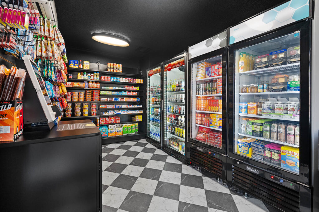 A convenience store interior with shelves stocked with snacks, canned goods, and drinks. Refrigerators on the right display beverages, dairy, and ready-to-eat foods. The floor has a black-and-white checkered pattern.