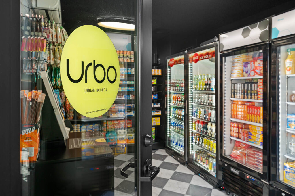 A view through a glass door labeled Urbo Urban Bodega into a well-lit convenience store with shelves and coolers stocked with snacks, drinks, and groceries. The floor has a black-and-white checkered pattern.