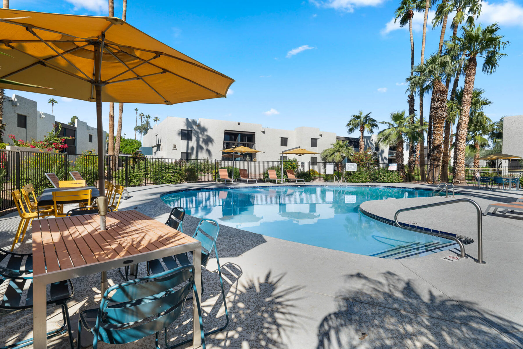 Outdoor swimming pool surrounded by lounge chairs, palm trees, and white apartment buildings under a blue sky; a shaded table with chairs and a yellow umbrella is in the foreground.
