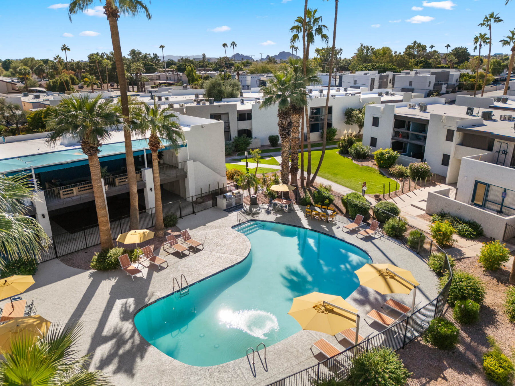 Aerial view of a modern apartment complex with a kidney-shaped outdoor pool, lounge chairs, and yellow umbrellas, surrounded by palm trees, green lawns, and white buildings under a clear blue sky.