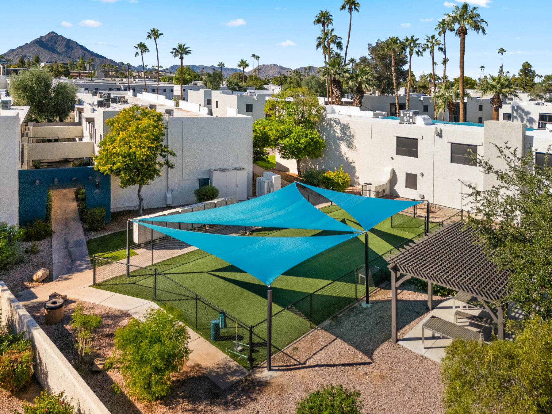 A fenced dog park with artificial turf, blue shade sails, benches, and a small pavilion, surrounded by white buildings, palm trees, and distant mountains under a sunny sky.