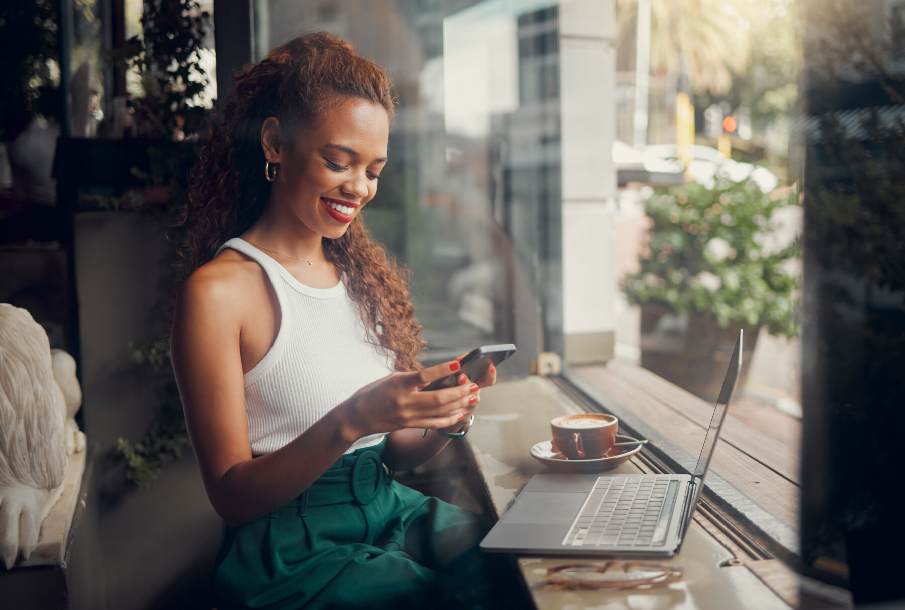 A woman sits by a window in a cafe, smiling at her phone. She wears a white sleeveless top and green pants, with a laptop and a cup of coffee on the table in front of her.